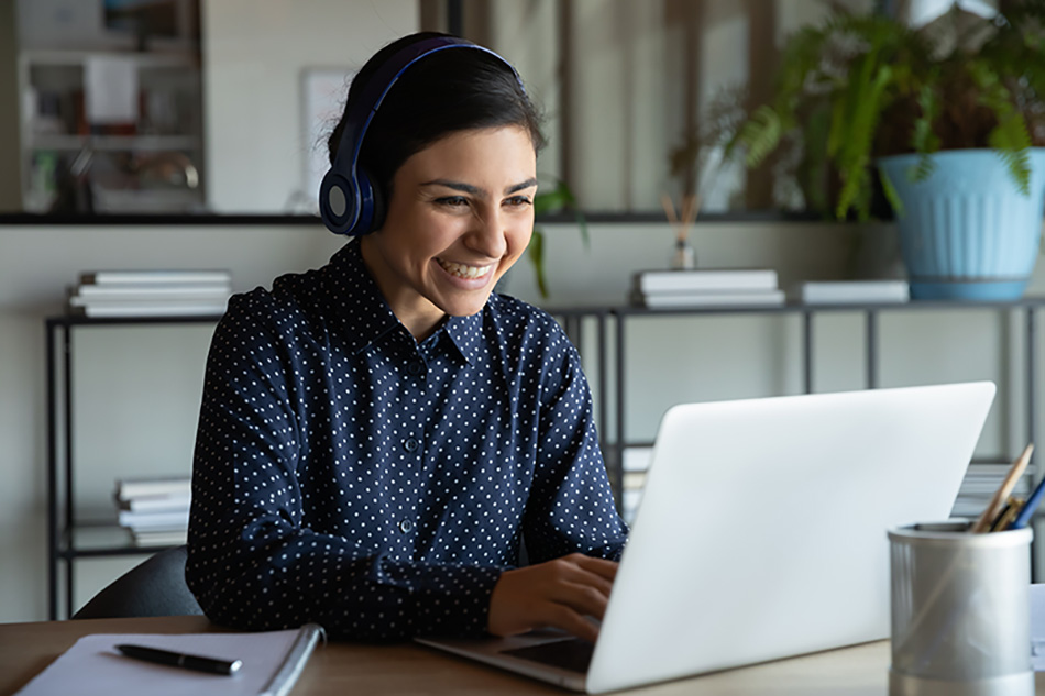 happy female wearing wireless headphones, looking at laptop screen, holding pleasant conversation with partners clients online, working remotely at workplace.