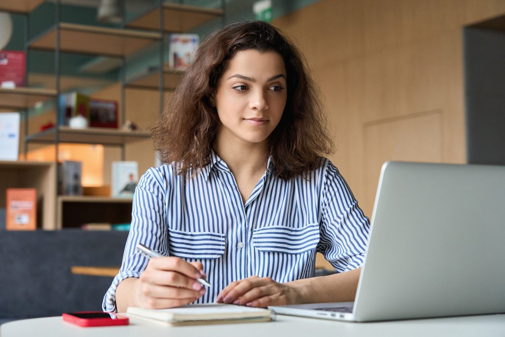 young woman writing in notepad while viewing her laptop computer 