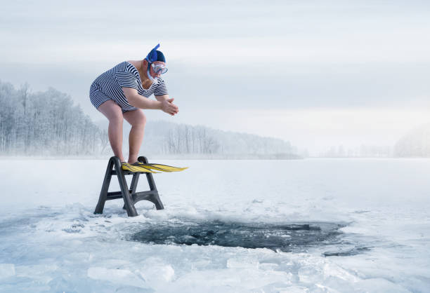 A man in a swimsuit standing on  step stool, outside in freezing temperatures, about to jump into a freezing pond. 