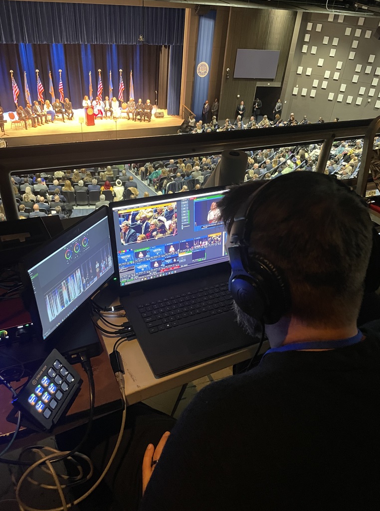 Looking over the shoulder of a man working a computer with multiple screens, with the inauguration ceremony happening on a stage in the background.