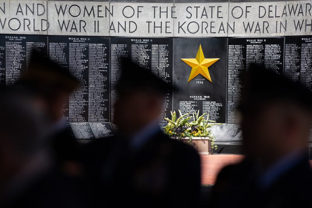 A memorial wall honoring the men and women of Delaware who served in World War II and the Korean War, featuring a prominent gold star and flower arrangements in front. Silhouettes of attendees are blurred in the foreground.