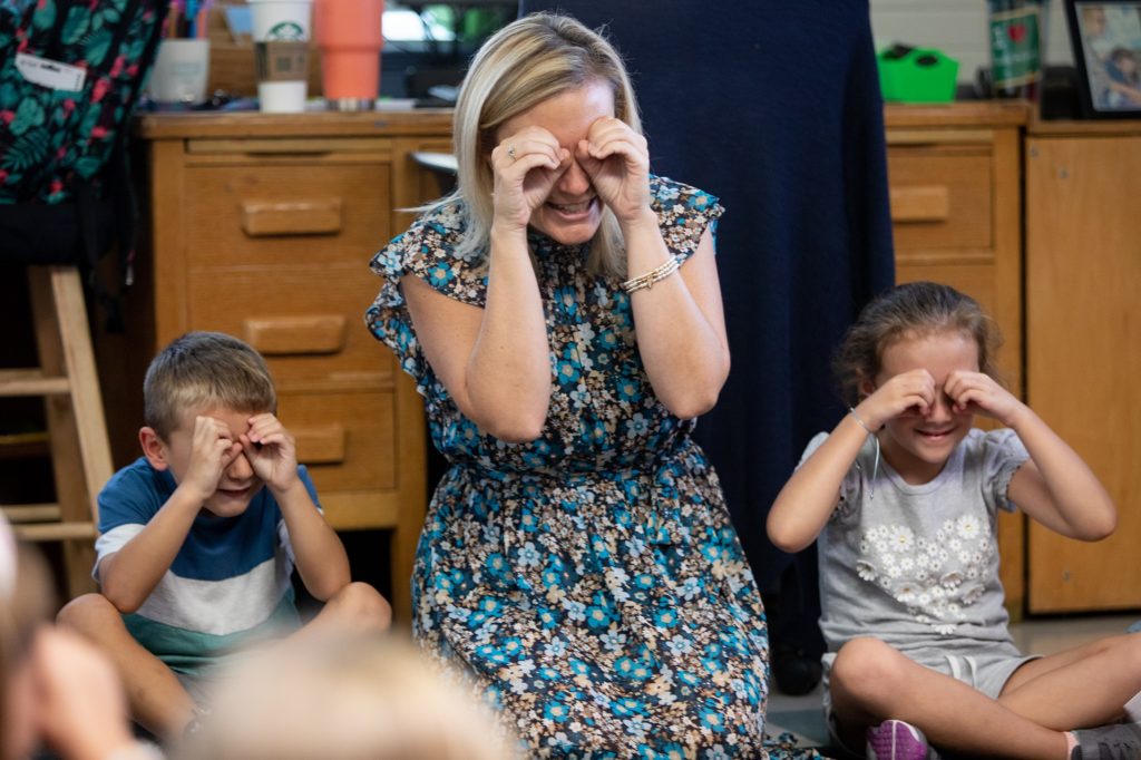 A teacher is sitting next to her students and they are all smiling. They have all put their hands up to their eyes like binoculars.
