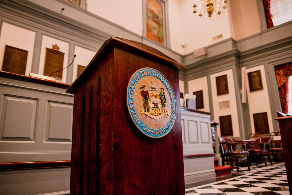 The podium in Legislative Hall in Dover, with the official state seal on the front. The room is lit but empty.