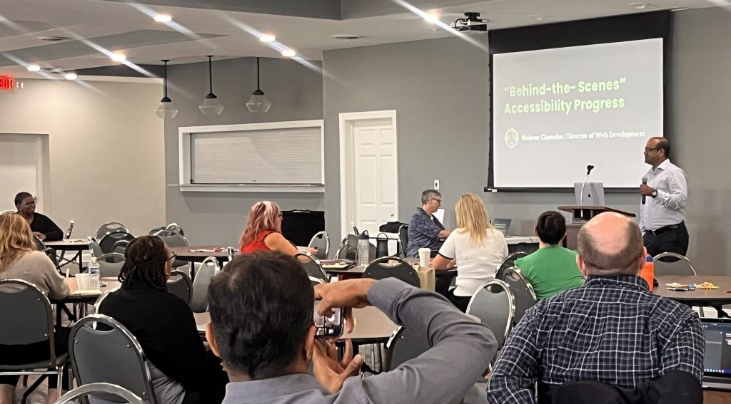 A conference room filled with people watching a man who is holding a microphone and speaking next to a screen.