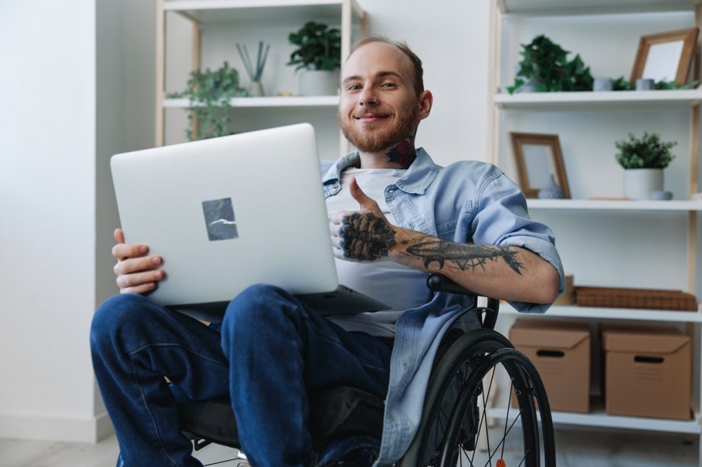 Man in wheelchair with laptop computer smiling and giving a thumbs-up sign. 